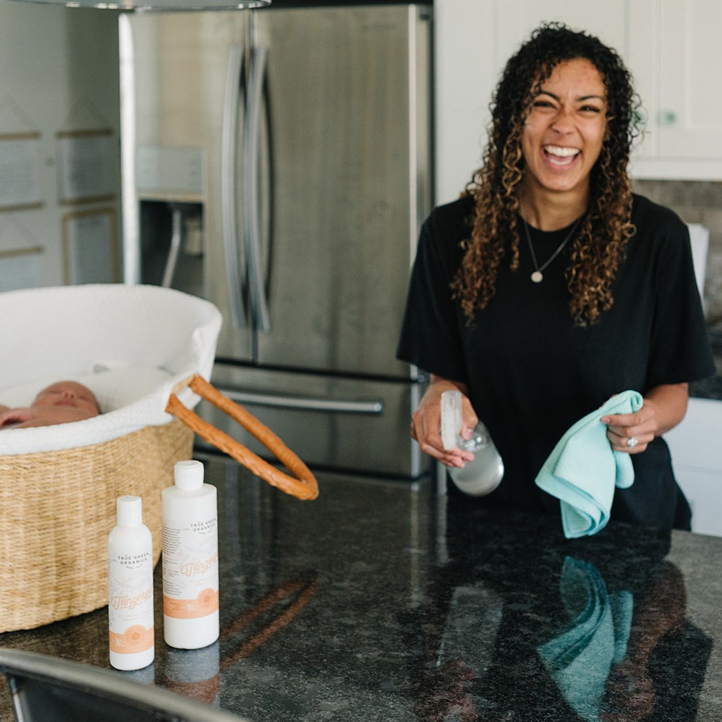 Woman smiling next to her baby in a bassinet cleaning counters with True Green Organics Tangerine Clean All Purpose Cleaner